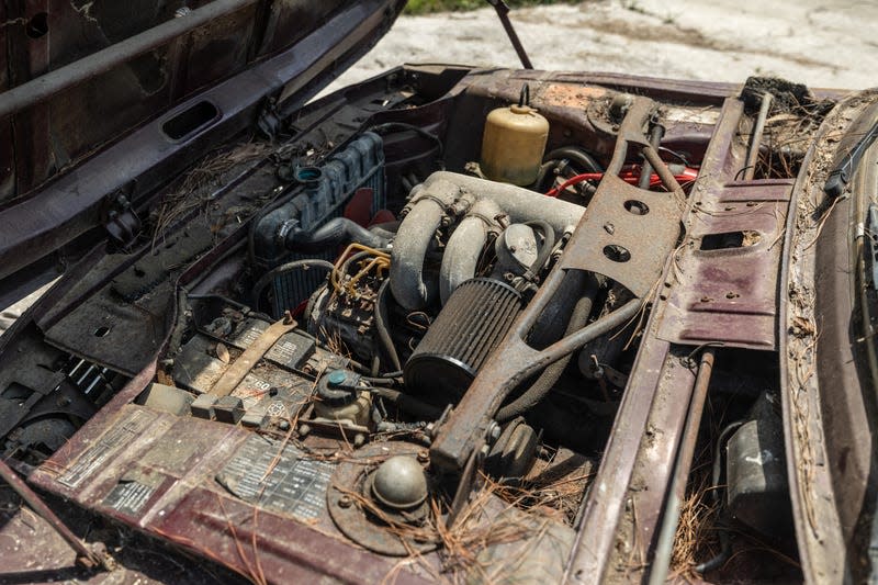 an under-hood photo of the engine bay of the burgundy 1974 bmw 2002tii. the car has sat abandoned in a field in florida for 25 years, and it shows: the engine compartment is full of leaves and branches and other trash, and the engine is covered in a thick layer of grime.