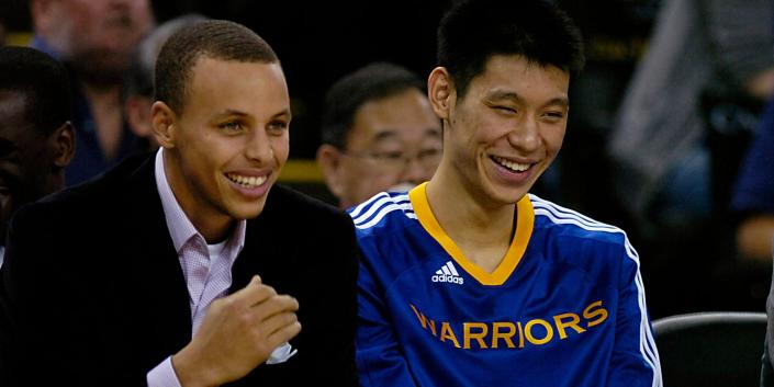 Stephen Curry, in a suit, laughs alongside Jeremy Lin, in Warriors warmups, while sitting on the bench in 2010.