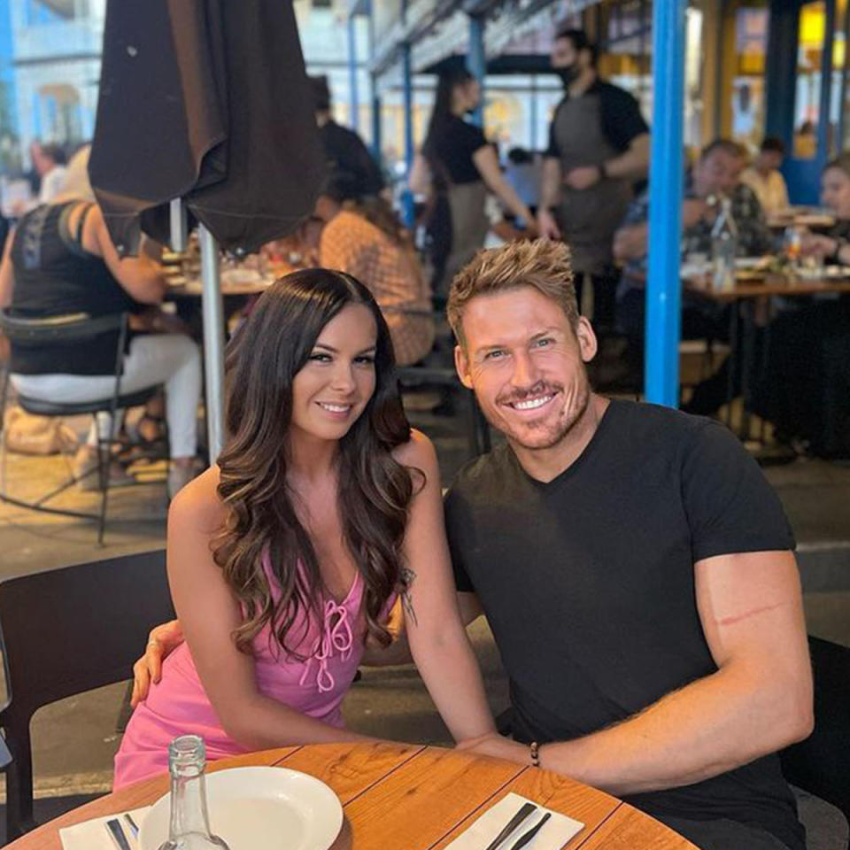 Seb Guilhaus in a black t-shirt poses with his girlfriend Ania who is wearing a pink dress. The pair are sitting at an outdoor dining area.