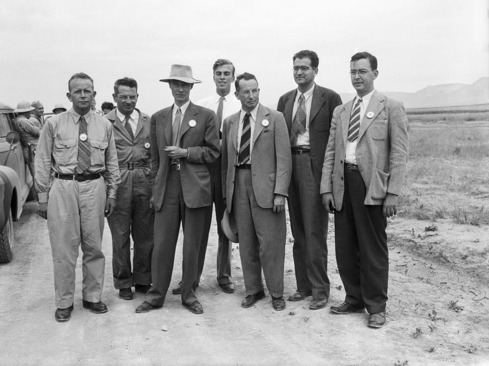 black and white photo shows seven men in suits standing on a dirt road in the new mexico desert