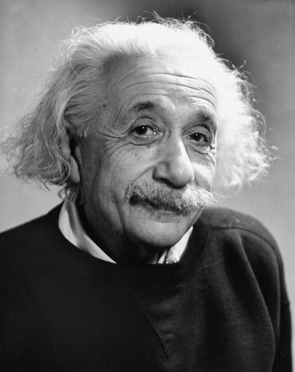 albert einstein looks at the camera, he wears a dark sweater over a collared shirt