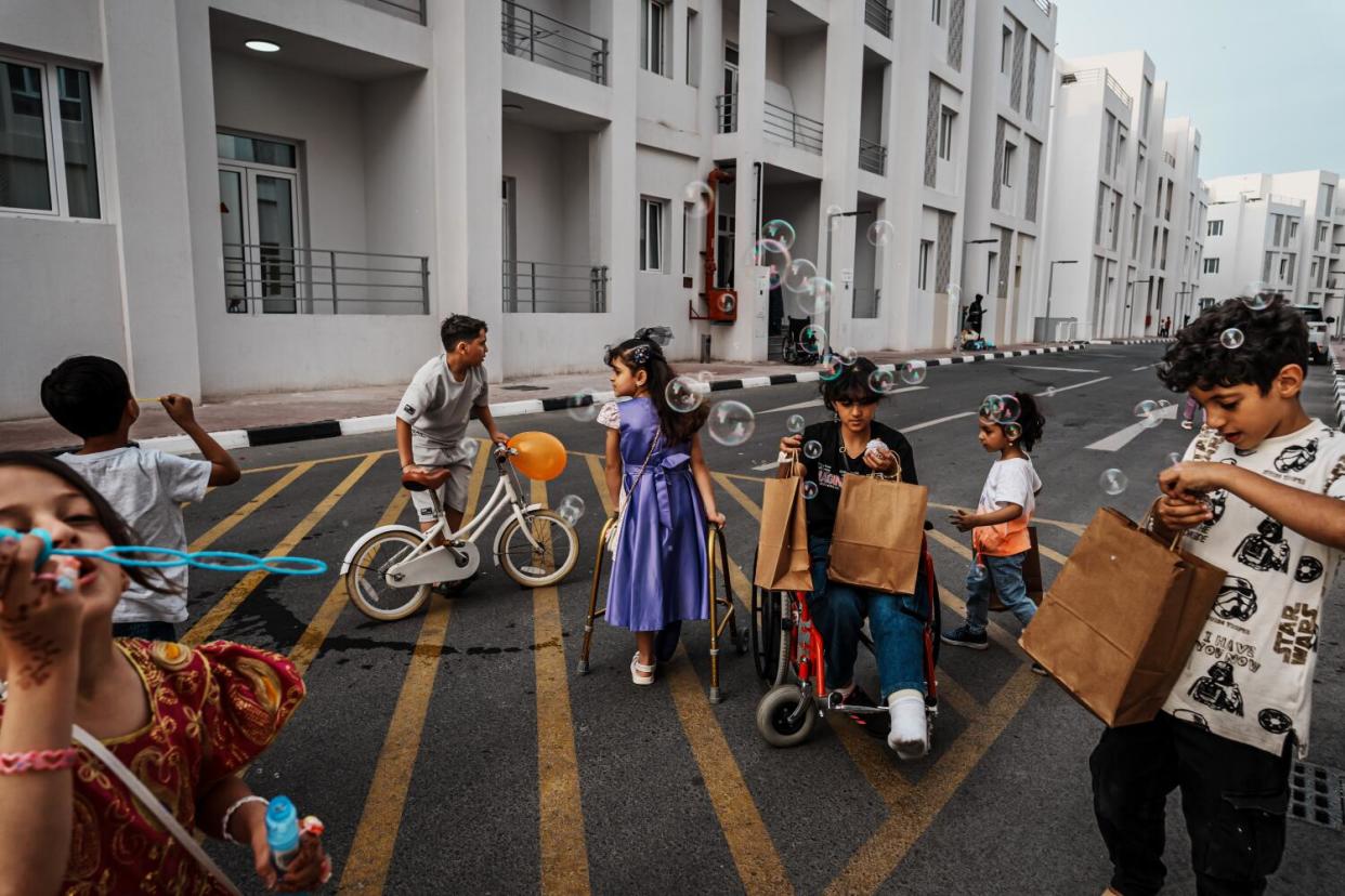 A group of children, including one in a wheelchair, holding bags, play on a street near a multistory building