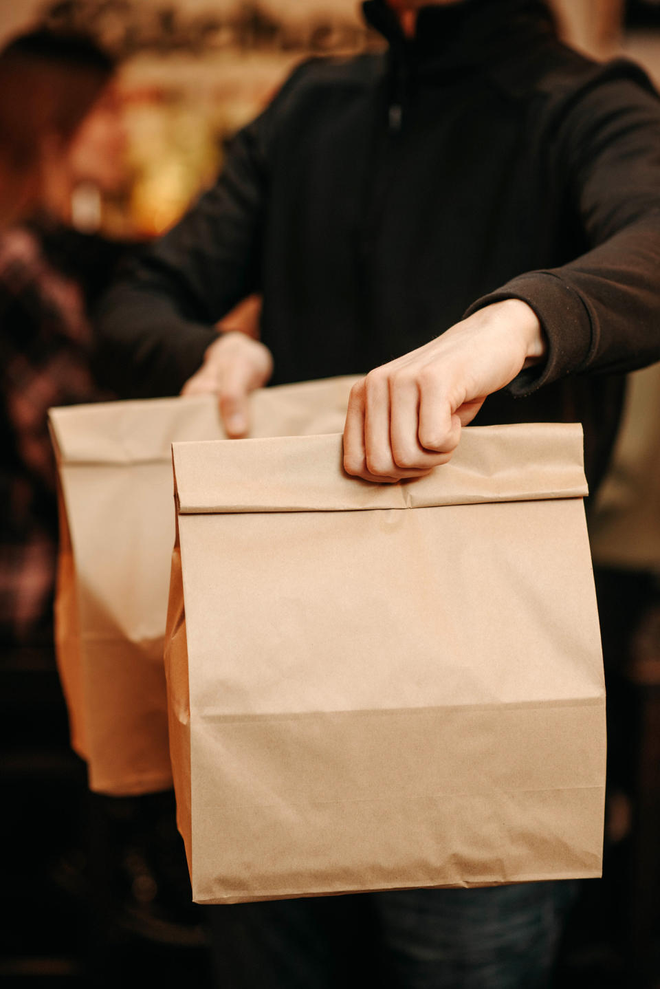 A person holding two brown paper bags with both hands, focus on the bags. Background is blurred, another person visible