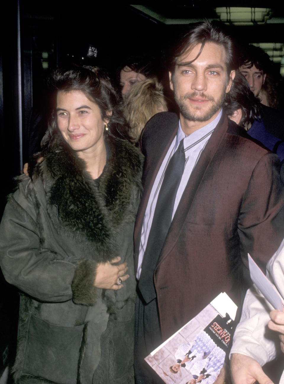 I don't know who these people are. A man in a suit and a woman in a fur-lined coat stand together, smiling at an event. The man is holding a book or program