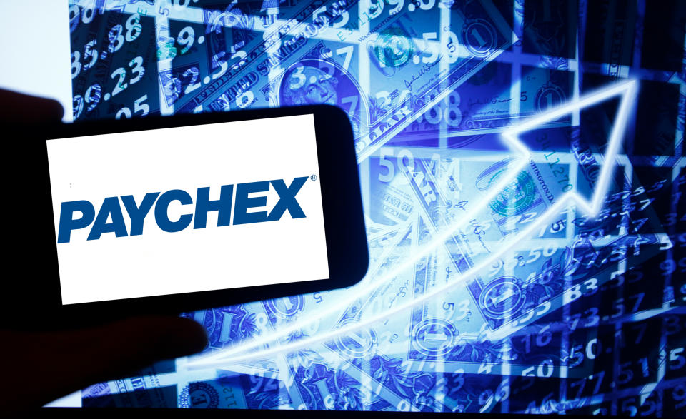 The logo of Paychex is seen on a screen of a smartphone next to a screen with an illustration of the stock market. Paychex is listed in Nasdaq. The Nasdaq is the second-largest stock exchange in the world after the New York Stock Exchange. (Photo by Alexander Pohl/NurPhoto via Getty Images)