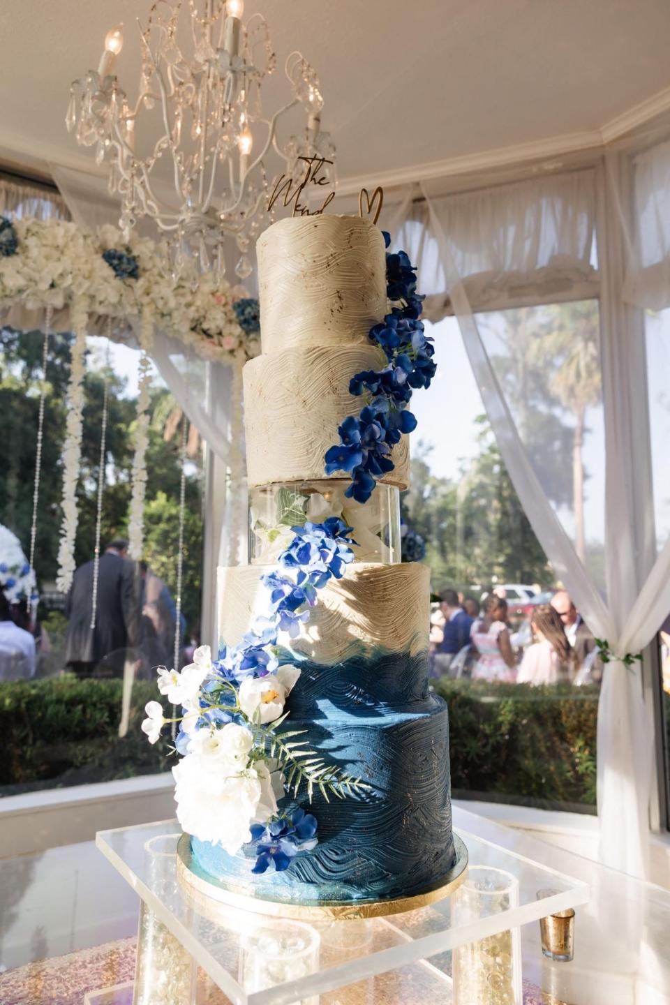 A tall, five-story wedding cake with floral detailing on a clear cake stand under a chandelier in a gazebo.