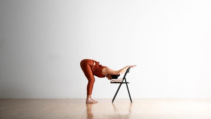 Woman practices Puppy Pose with a chair. She places her head on a folded blanket in the seat of a folding chair. Her arms reach over the back of the seat.