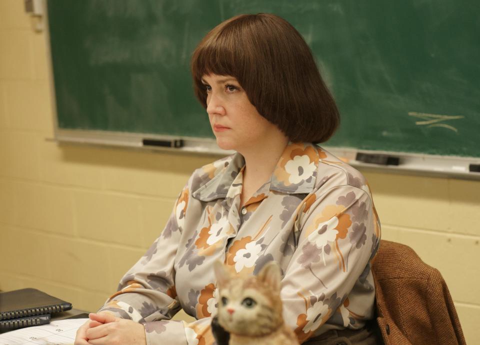 Woman in a floral blouse sits at a desk, with a small figure on the table, in a classroom setting