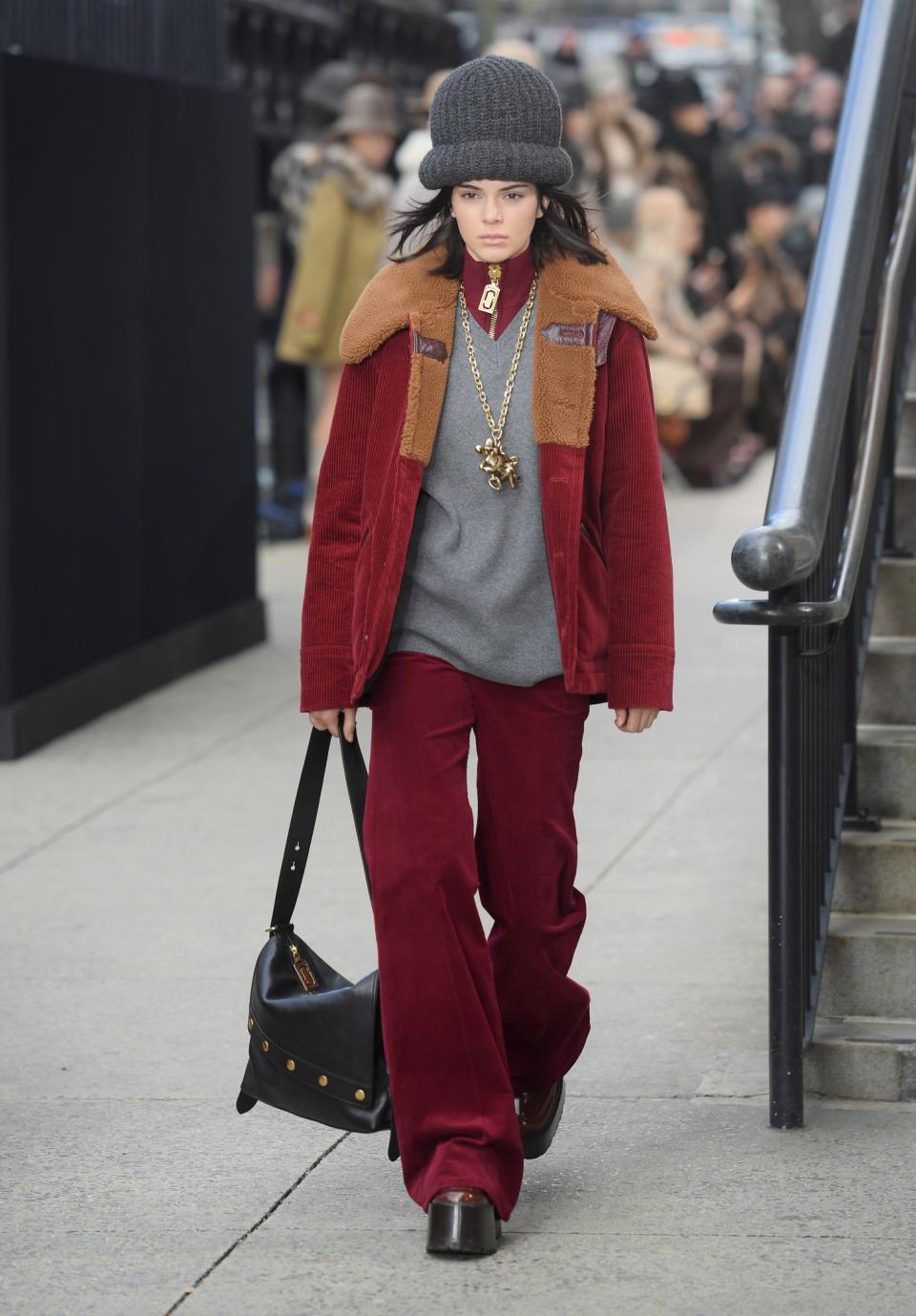 Model Kendall Jenner wears a large bowling hat, burgundy red corduroy jacket, matching velvet pants, and large gold chain necklace