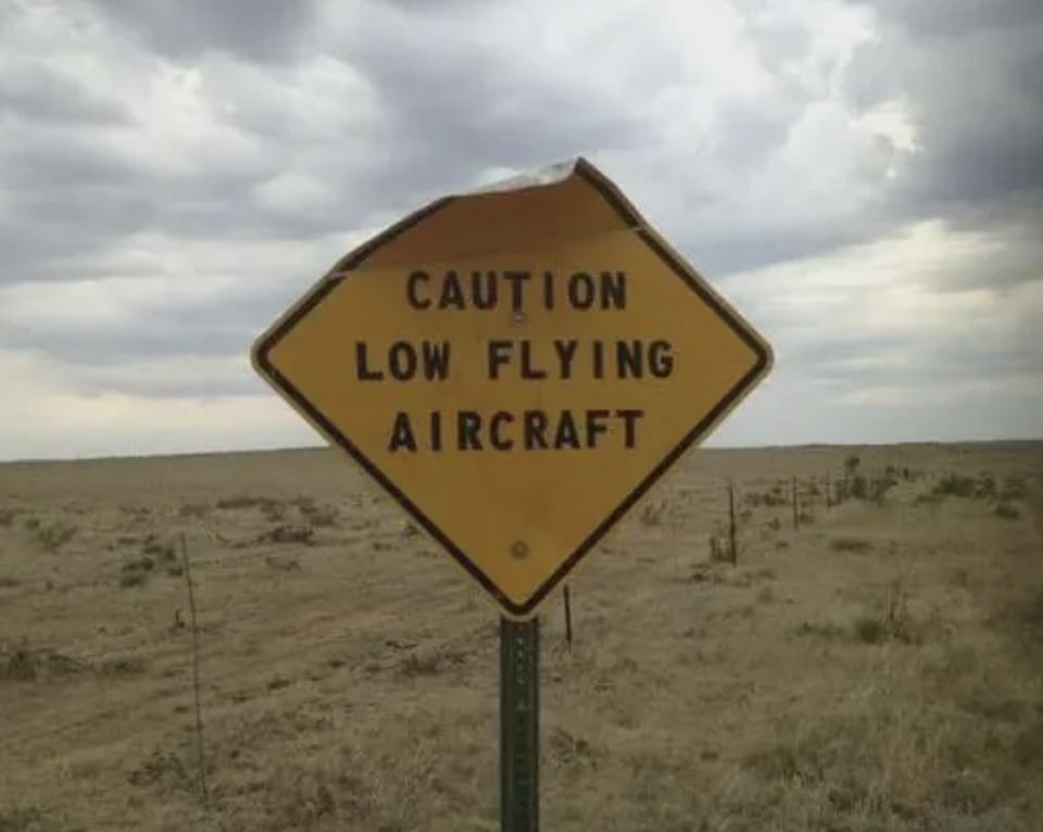 Warning sign reads "CAUTION LOW FLYING AIRCRAFT" with a flat landscape in the background