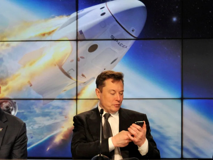 Elon Musk looks at his phone with a display of a space capsule entering orbit behind him.