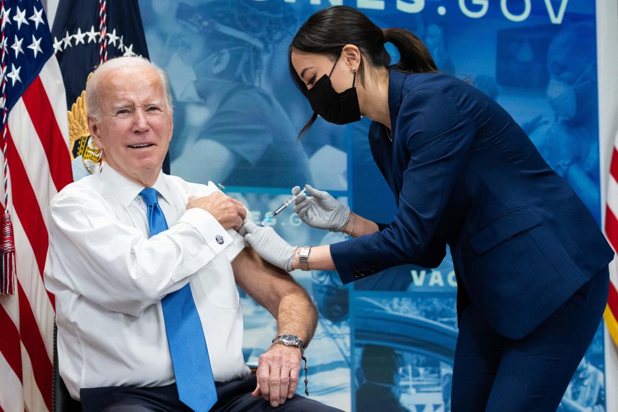 President Biden, with a sleeve rolled up, gets a shot in the arm by a woman wearing rubber gloves and a face mask.