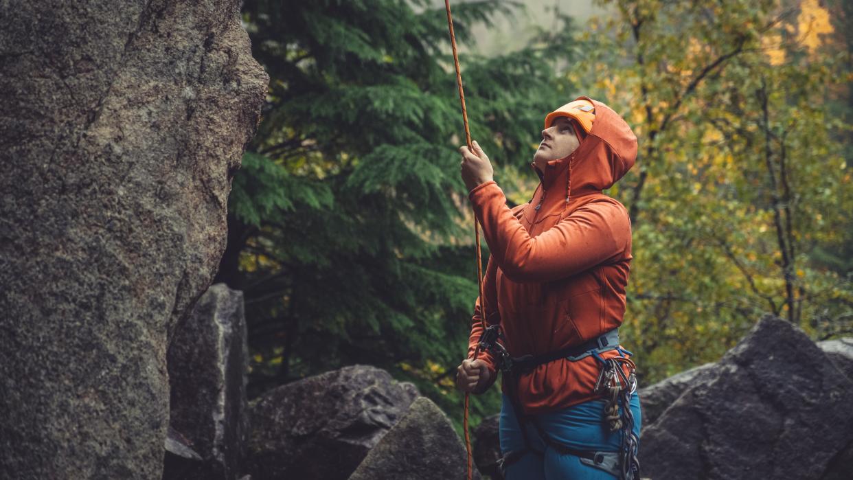  A rock climber belays at the bottom of a cliff surrounded by trees with autumn coloured leafs . 