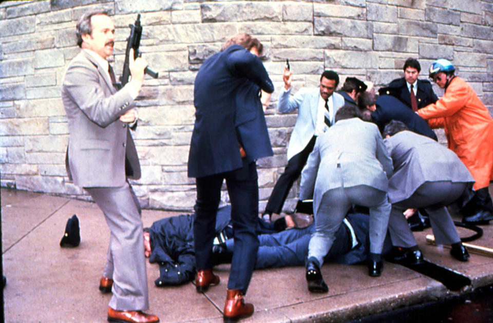 Secret Service agents with guns drawn swarm around Thomas Delahanty and James Brady, both in prone position, while apprehending shooting suspect John Hinckley Jr.