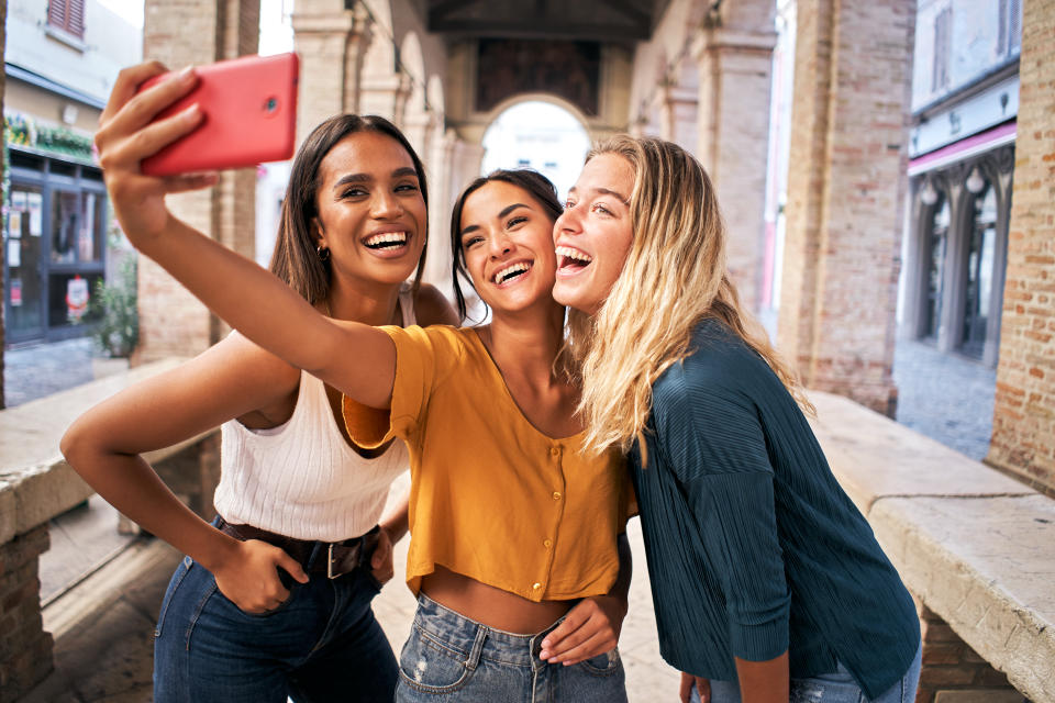 Tiga pacar ceria dengan pakaian musim panas berfoto selfie di luar ruangan di tengah kota turis.  Gambar berkualitas tinggi