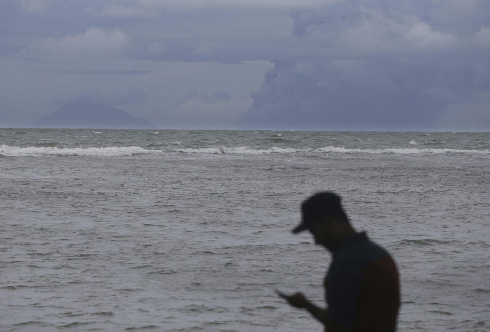 A man look a mobile phone as Anak Krakatau, right, is seen on the horizon off Carita beach, Indonesia, Friday, Dec. 28, 2018. Indonesia raised the danger level for an island volcano that triggered a tsunami on the weekend, killing at least 430 people in Sumatra and Java, and widened its no-go zone.(AP Photo/Achmad Ibrahim)