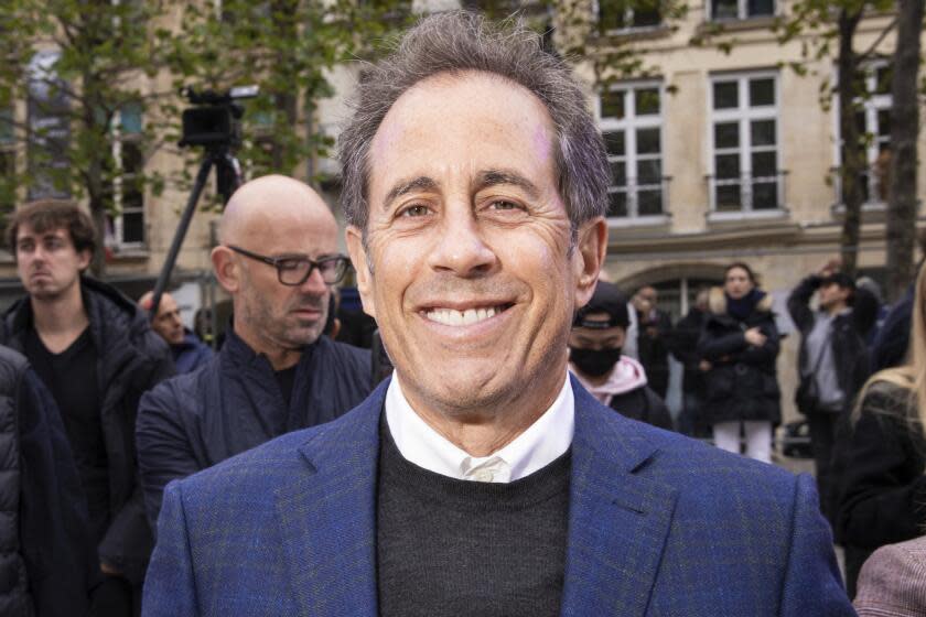Jerry Seinfeld in a dark blue blazer, black sweater and white dress shirt smiling in the middle of a crowd