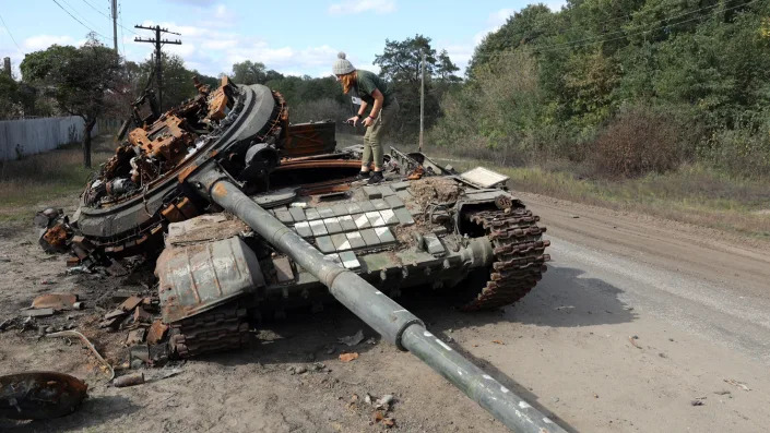 A person stands on top of a destroyed tank near the side of a road.