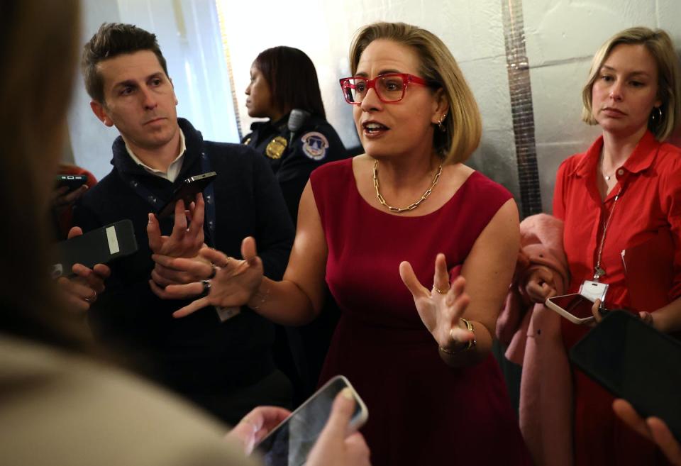 Kyrsten Sinema wears a red dress and red framed glasses and gestures with her hands, while people stand around her and hold out phones and tape recorders.