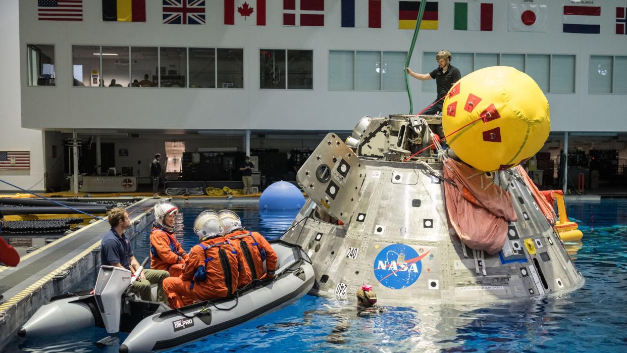  A cone-shaped capsule in a pool with a raft beside it. the raft has 3 astronauts in orange pumpkin suits . 