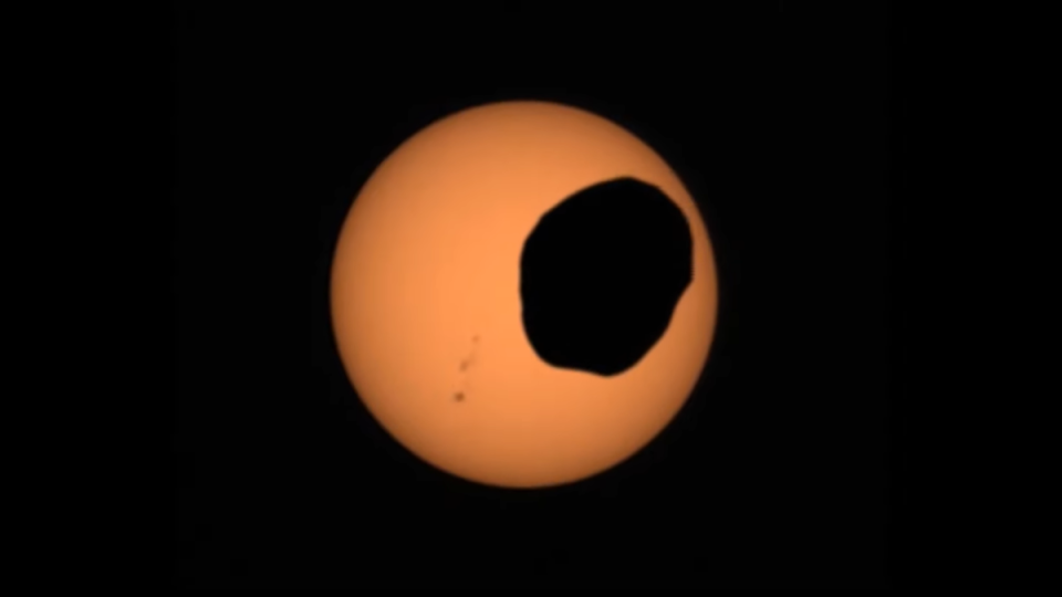 A screenshot from a video showing the Mars moon Phobos crossing the face of the sun. NASA’s Perseverance Mars rover captured the video on April 2, 2022, with its Mastcam-Z camera. You can also spot a group of sunspots on the left.