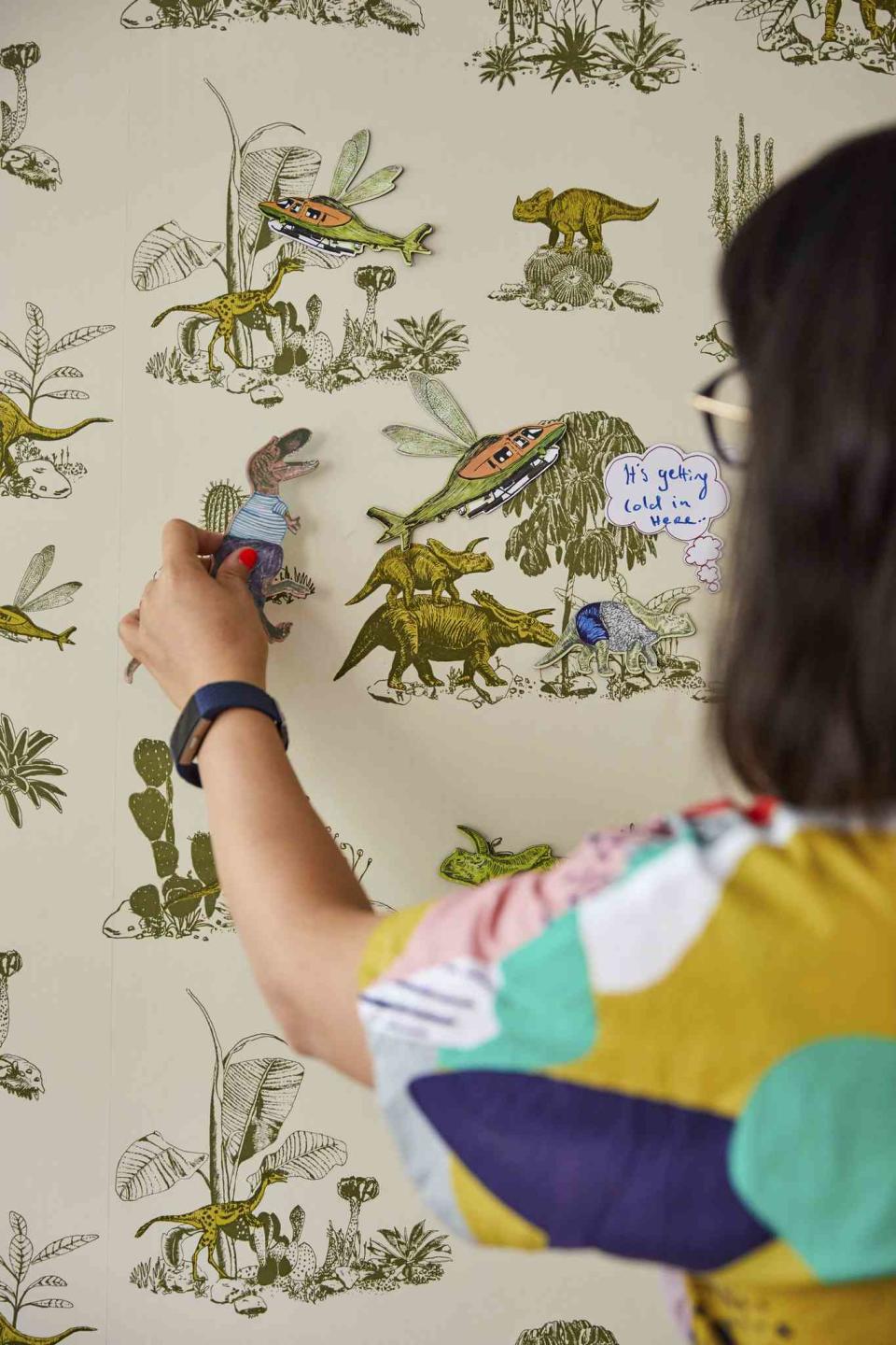 Sian Zeng standing in front of a wall decorated with green and yellow print wallpaper, holding a dinosaur figure up to the wall