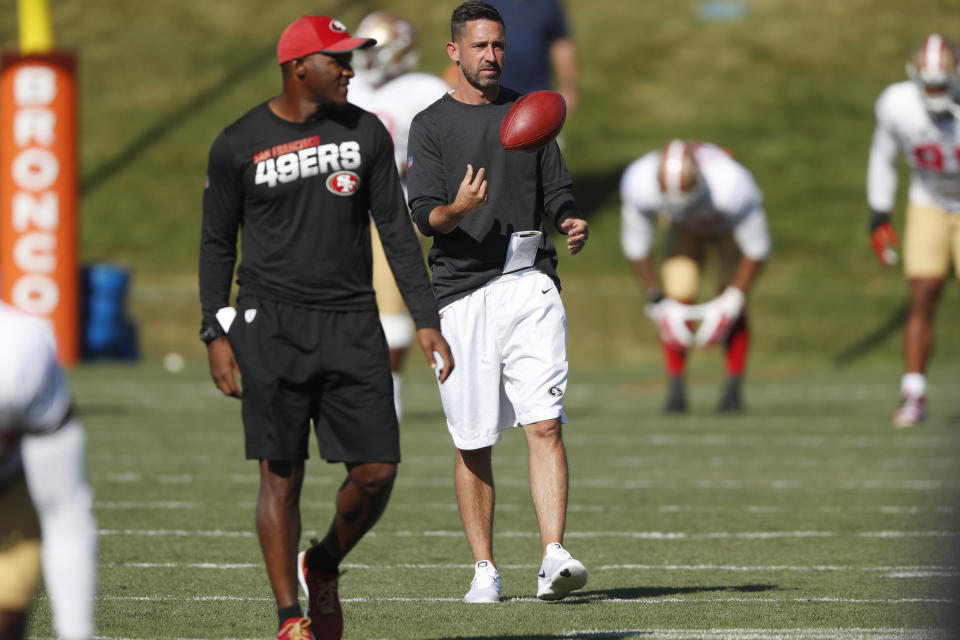 San Francisco 49ers head coach Kyle Shanahan, back, tosses a ball as defensive backs coach Joe Woods looks on during a combined NFL training camp with the Denver Broncos Saturday, Aug. 17, 2019, at the Broncos' headquarters in Englewood, Colo. (AP Photo/David Zalubowski)