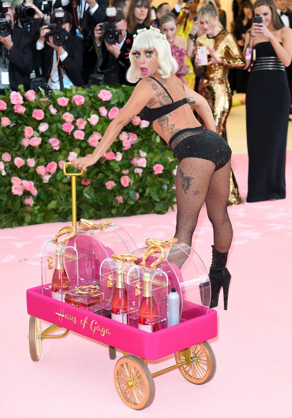 Lady Gaga poses on the Met Gala red carpet in a mesh lingerie outfit and pulling a pink wagon full of alcohol that says "Haus of Gaga" on the side.