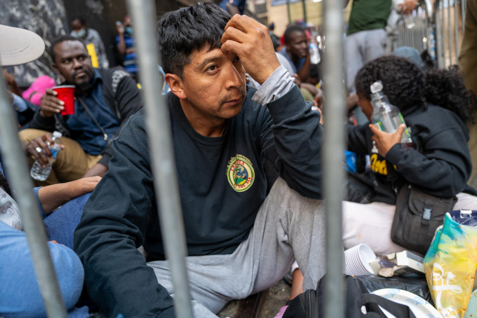 A migrant, looking drawn and tired, sits on the sidewalk with many others behind a barrier outside the Roosevelt Hotel.