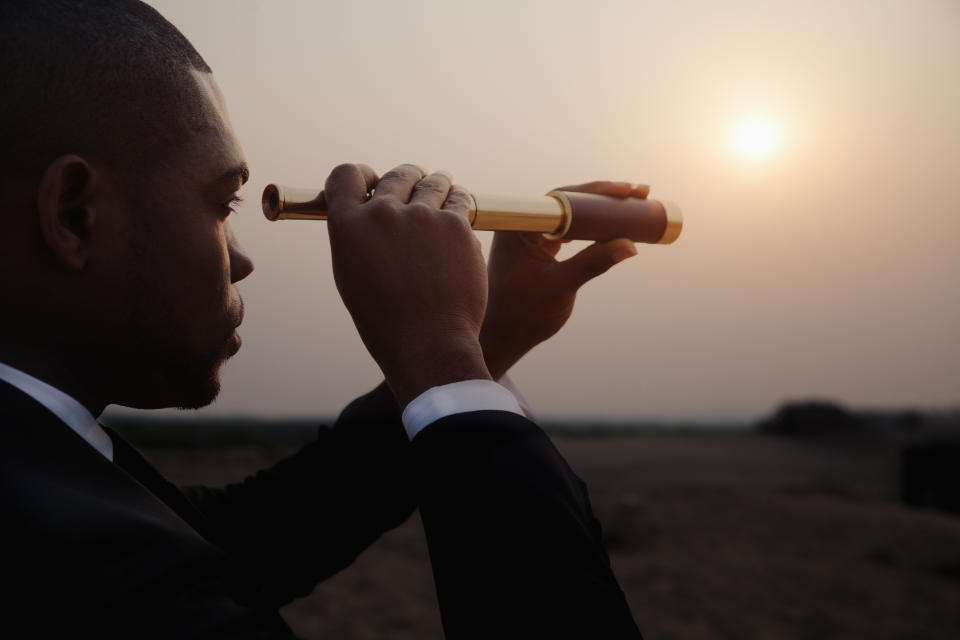 A man in a suit looks through a spyglass in the desert.