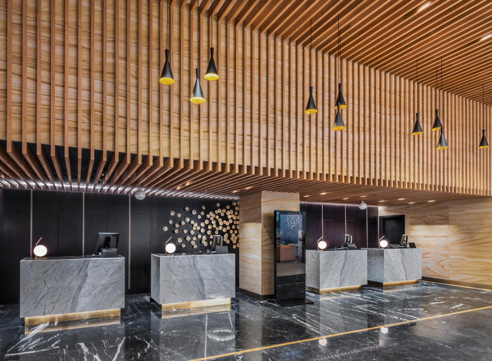 A modern hotel lobby with marble floors, four check-in desks with dark stone finishes, and a wooden slat ceiling with pendant lights hanging above