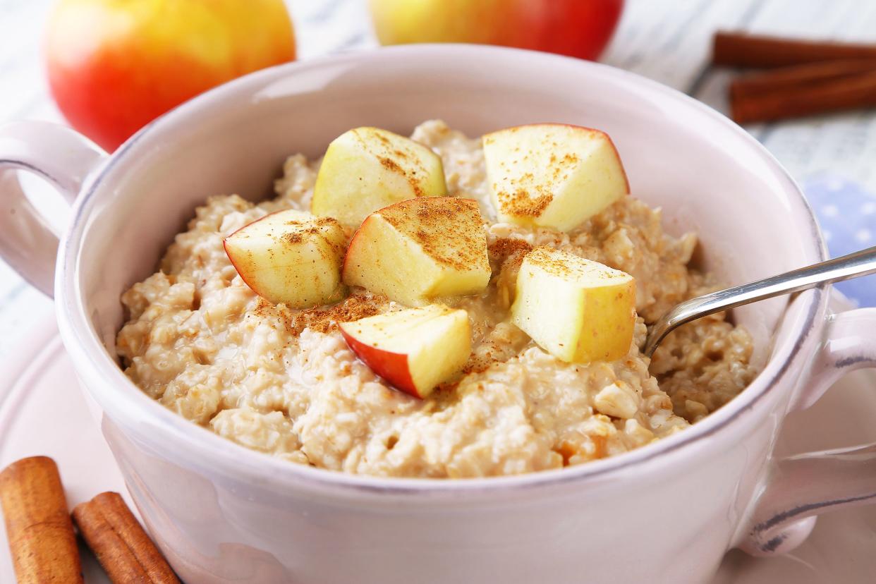 Closeup of apple cinnamon oatmeal with a spoon in a white bowl, surrounded by ingredients, with a blurred background