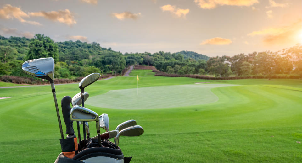 Golf course at sunset with a set of golf clubs in the foreground. The green is well-maintained with a flag visible in the distance