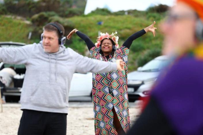 A woman wearing a colourful outsift smiling widely with her arms stretched out. She is outside on what looks like a beach.