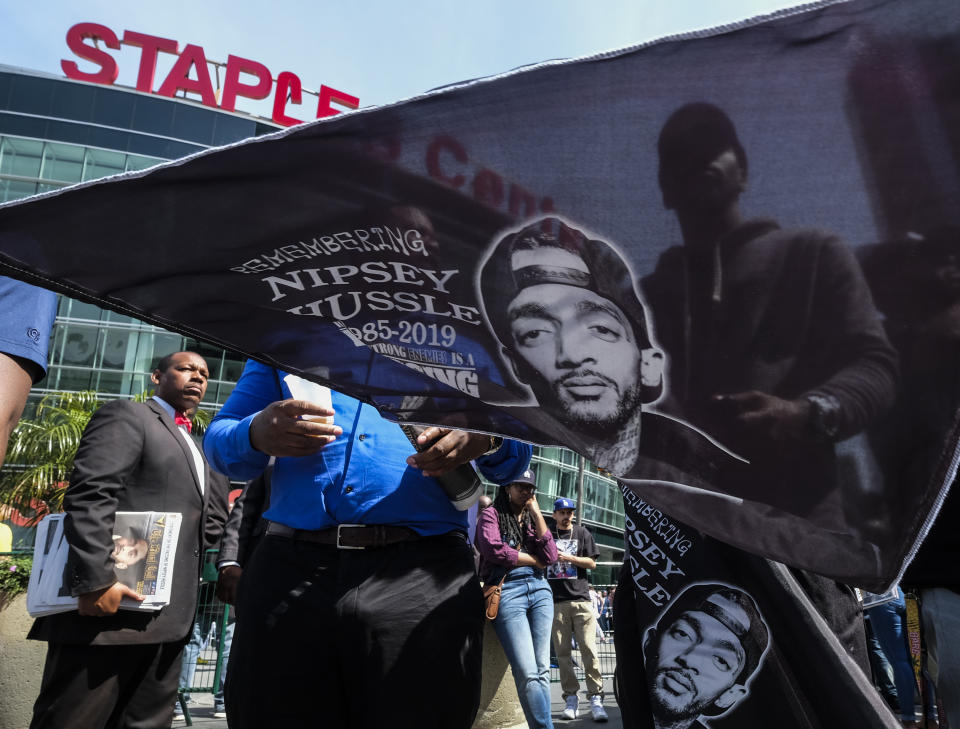 Un fan del difunto Nipsey Hussle ondea una bandera con la imagen del rapero durante un servicio memorial público en el Staples Center en Los Ángeles, el jueves 11 de abril del 2019. (AP Foto/Ringo H.W. Chiu)