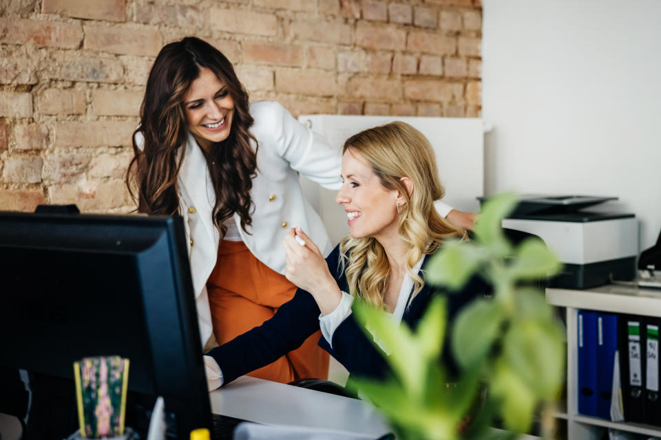 Two women smiling and working together at a desk in an office setting with a computer and office supplies around