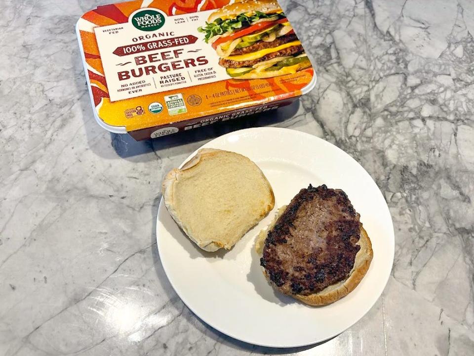 An orange box with a Whole Foods label and an image of a burger on it next to a white plate with a burger patty on a bun and the top of the bun placed on the side