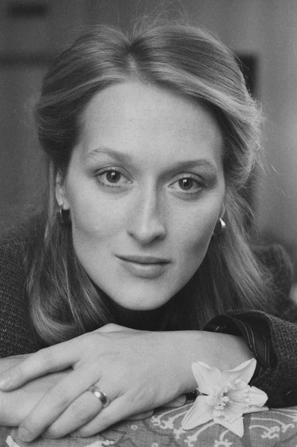 Meryl Streep posing with a neutral expression, her hands resting on a surface, adorned with a flower. She is wearing a simple outfit and hoop earrings