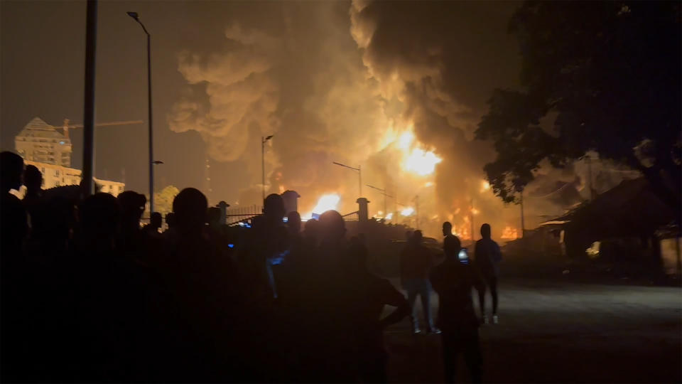 In this image made from video, residents watch a plume of smoke from a burning oil depot, in Conakry, Guinea, Monday, Dec. 18, 2023. An explosion and inferno at Guinea’s main fuel depot in the capital of Conakry has left several people dead or injured. Guinea’s presidency says the fire broke out at the Guinean Petroleum Company depot shortly after a massive explosion past midnight Sunday. (AP Photo)