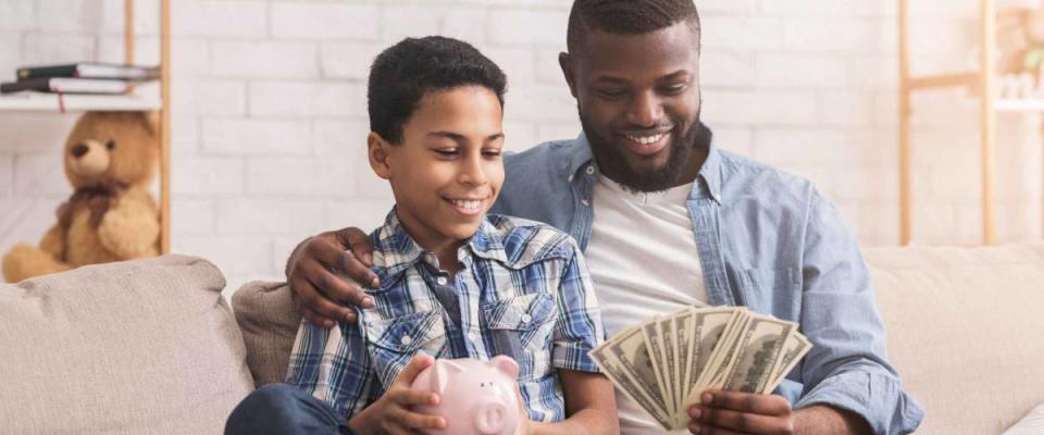 Father and son sitting on a couch, smiling, holding cash and a pink piggy bank