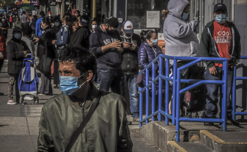 A masked pedestrian waits to cross a street after walking past a line of people in Brooklyn's Sunset Park—a neighborhood with one of the city's largest Mexican and Hispanic community, wearing masks to help stop the spread of coronavirus while waiting in line to enter a store, Tuesday May 5, 2020, in New York. A poll found that 61% of Hispanic Americans say they've experienced some kind of household income loss as a result of the COVID-19 outbreak. (AP Photo/Bebeto Matthews)