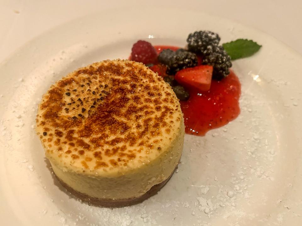 A small cheesecake with caramelized sugar on top. The cheesecake is on a white plate with a small side of berries in a syrup.