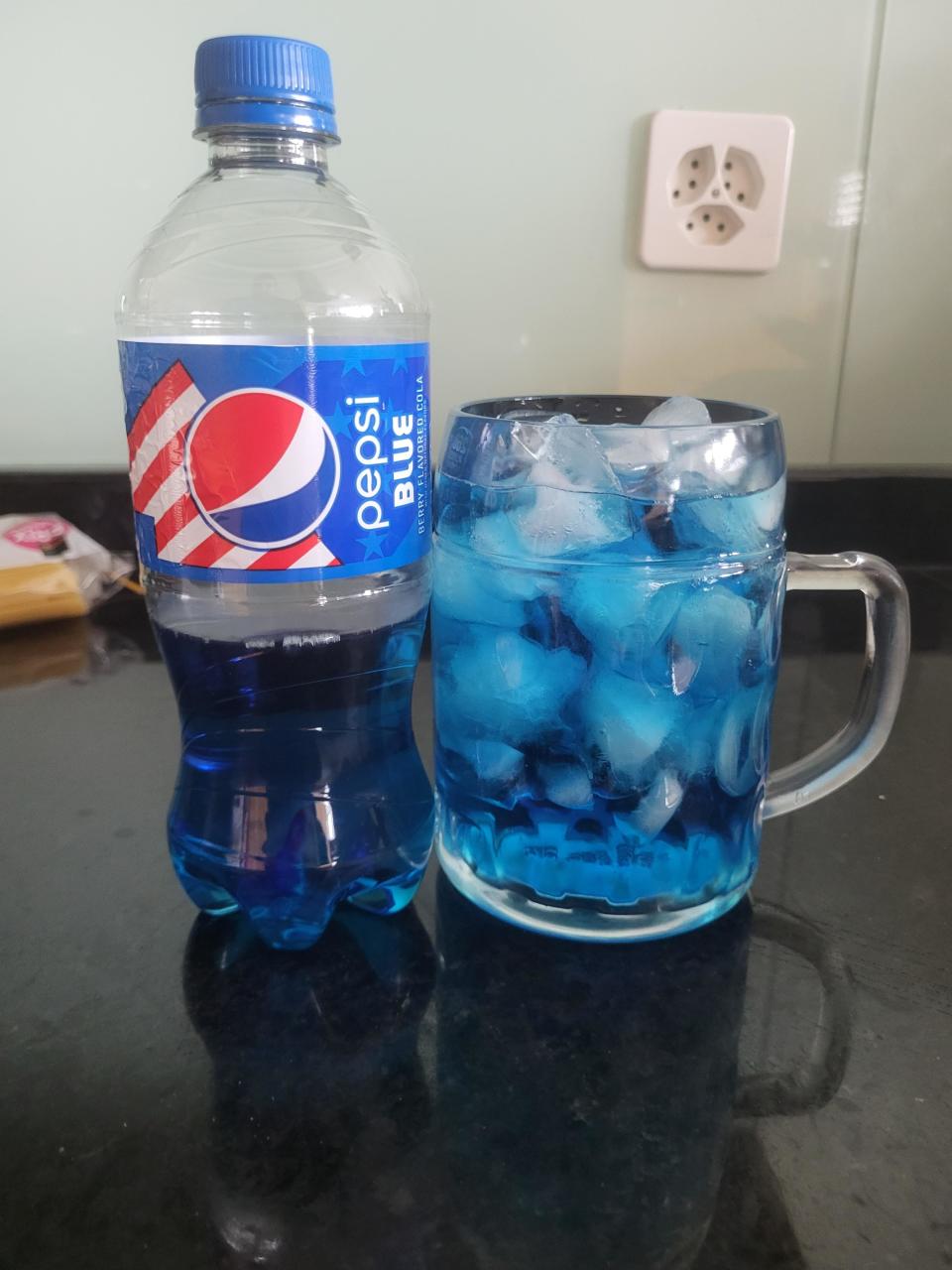 A bottle of Pepsi Blue next to a glass mug filled with ice and blue liquid. An electrical outlet is visible in the background