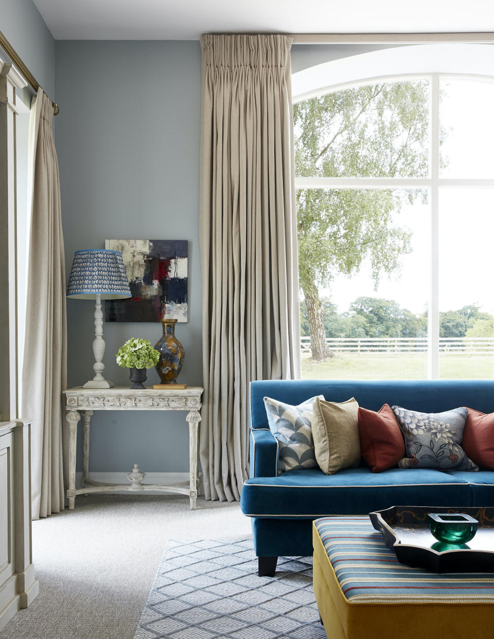 living room with blue couch, cream drapes, console, table lamps, patterned cushions, footstool