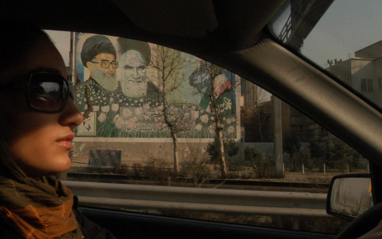 A young woman wearing a loose veil and sunglasses drives past a mural of Iranian leaders Ayatollah Khomeini (right) and Ayatollah Khamenei, on the Modaress highway in Tehran, Iran, 19th January 2006. (Photo by Kaveh Kazemi/Getty Images) - Kaveh Kazemi/Getty Images