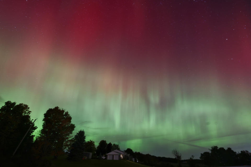 A night sky with aurora borealis displaying vibrant patterns above a rural landscape with trees and a small house