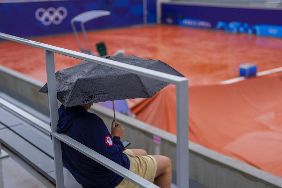 Un espectador con un paraguas sentado bajo la lluvia de una de las canchas de Roland Garros. Una decena de partidos fueron demorados a causa del mal tiempo en la primera jornada del tenis en los Juegos Olímpicos de París, sábado 27 de julio de 2024. (AP Foto/Manu Fernández)