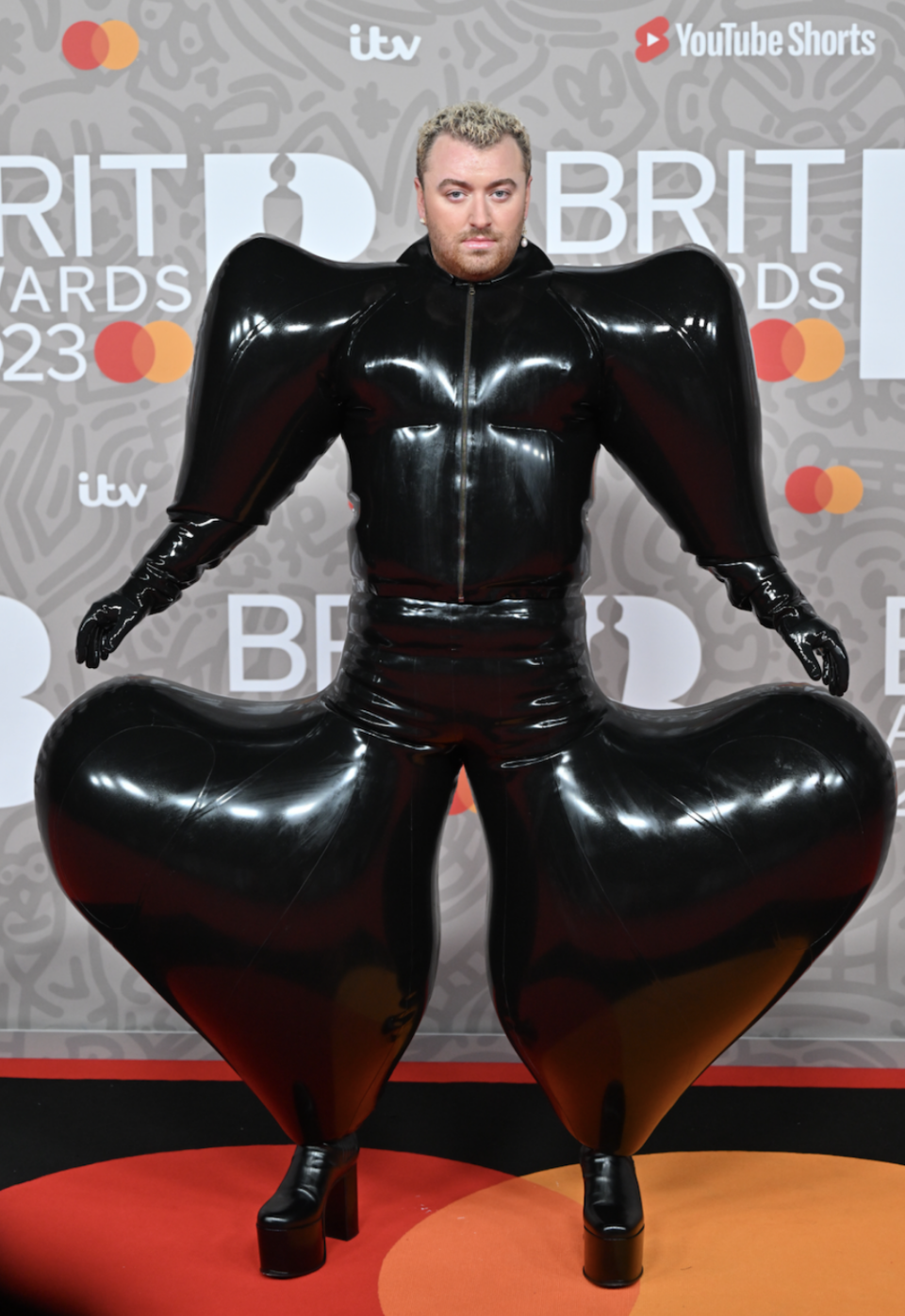 sam in a latex suit with exaggerated shoulders and pants coming out in a large heart shaped curve