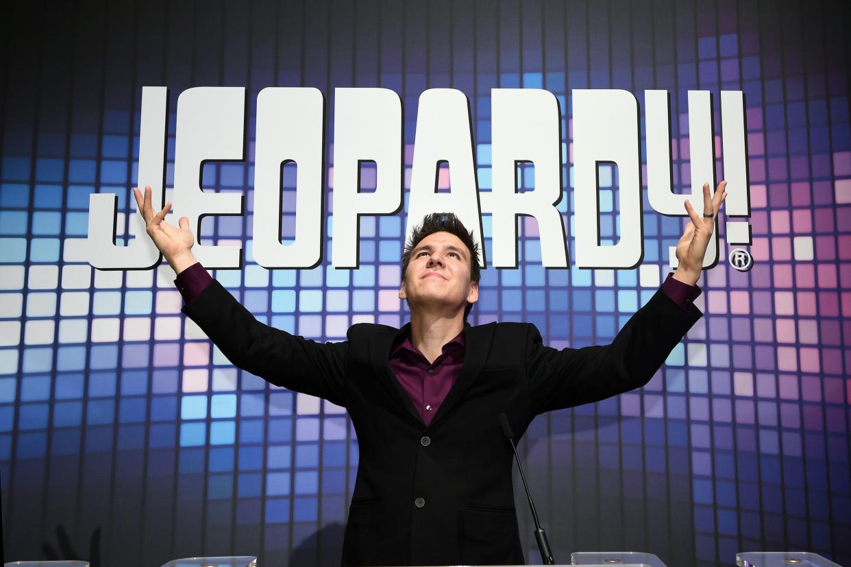 James Holzhauer poses in front of a Jeopardy sign with his arms raised