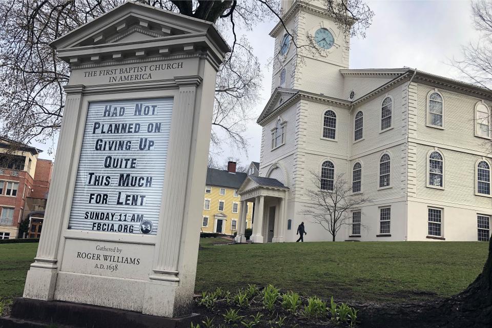 In this March 24, 2020, photo, a man walks past the First Baptist Church in America in Providence, R.I. Americans are turning to humor in many forms, like the sign in front of the church, as they cope with the fear and anxiety the coronavirus pandemic has unleashed. (AP Photo/William J. Kole)